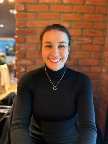 Woman in black wearing necklace with circle on it smiles in front of brick wall