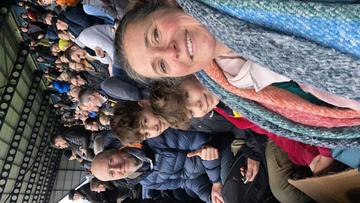 Louise sits in the stands at a football match. She has brown hair and wearing a multicoloured scarf. Two young boys smile at the camera behind her, and so does an man behind them. 